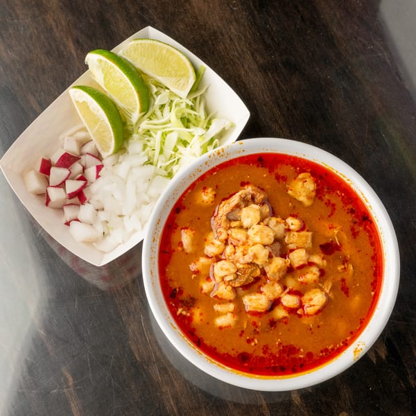 A bowl of pozole with a side of diced radishes, onions, shredded cabbage, and lime wedges.