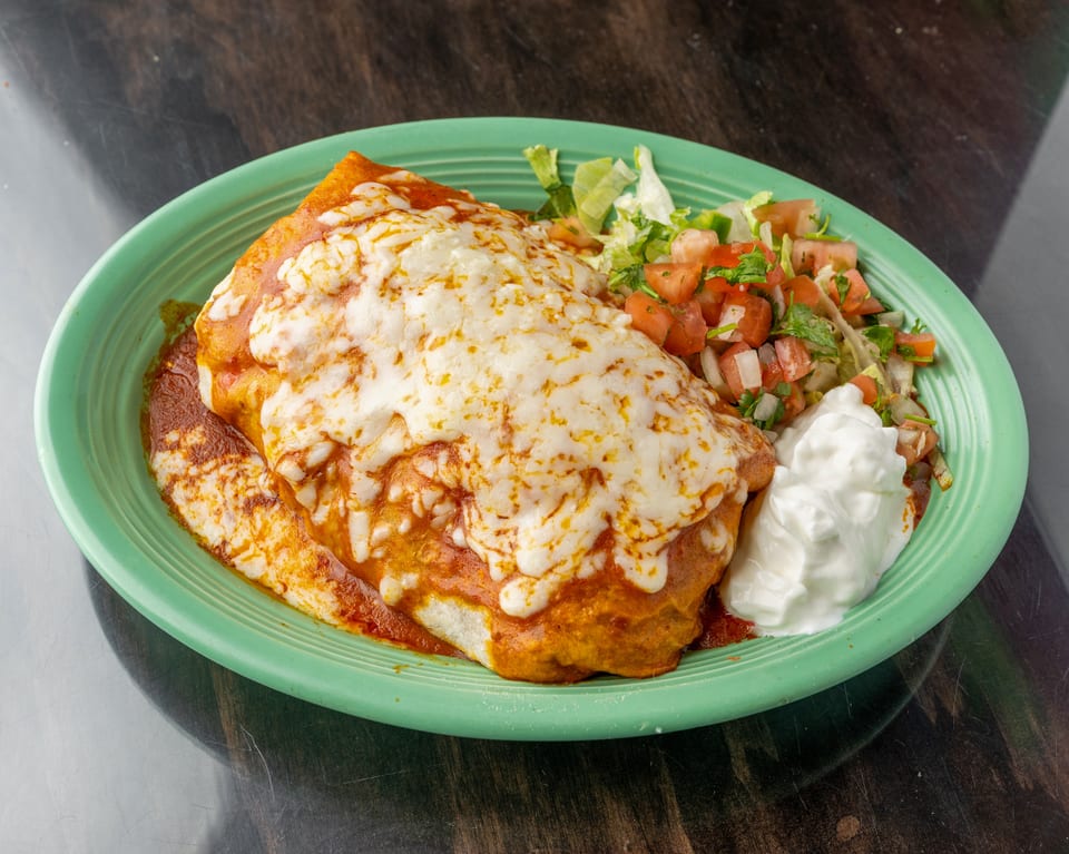 A large, cheesy burrito covered in red sauce, served with pico de gallo and sour cream.