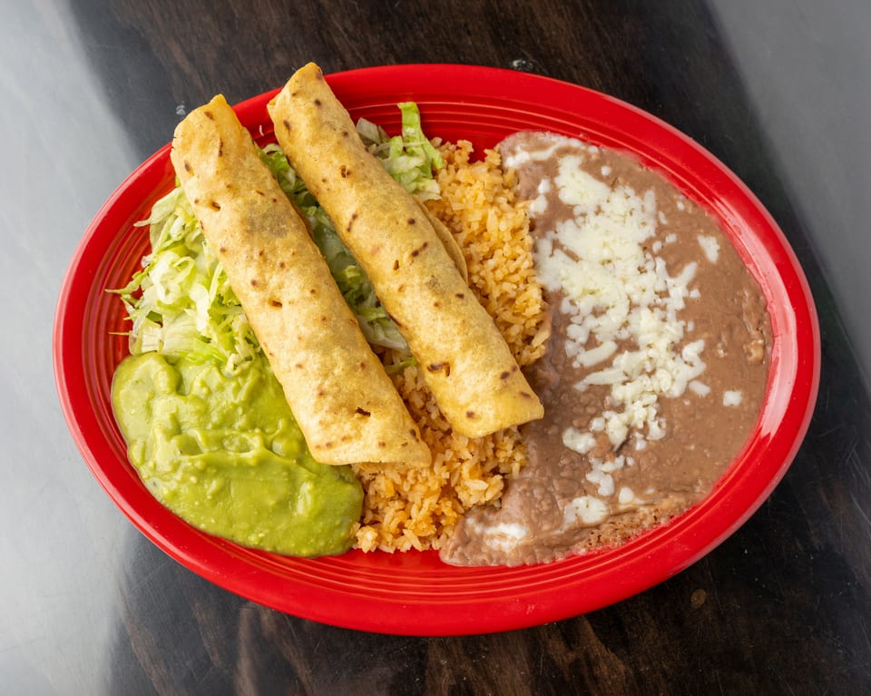 Two golden taquitos with shredded lettuce, guacamole, rice, and refried beans with cheese on a red plate.