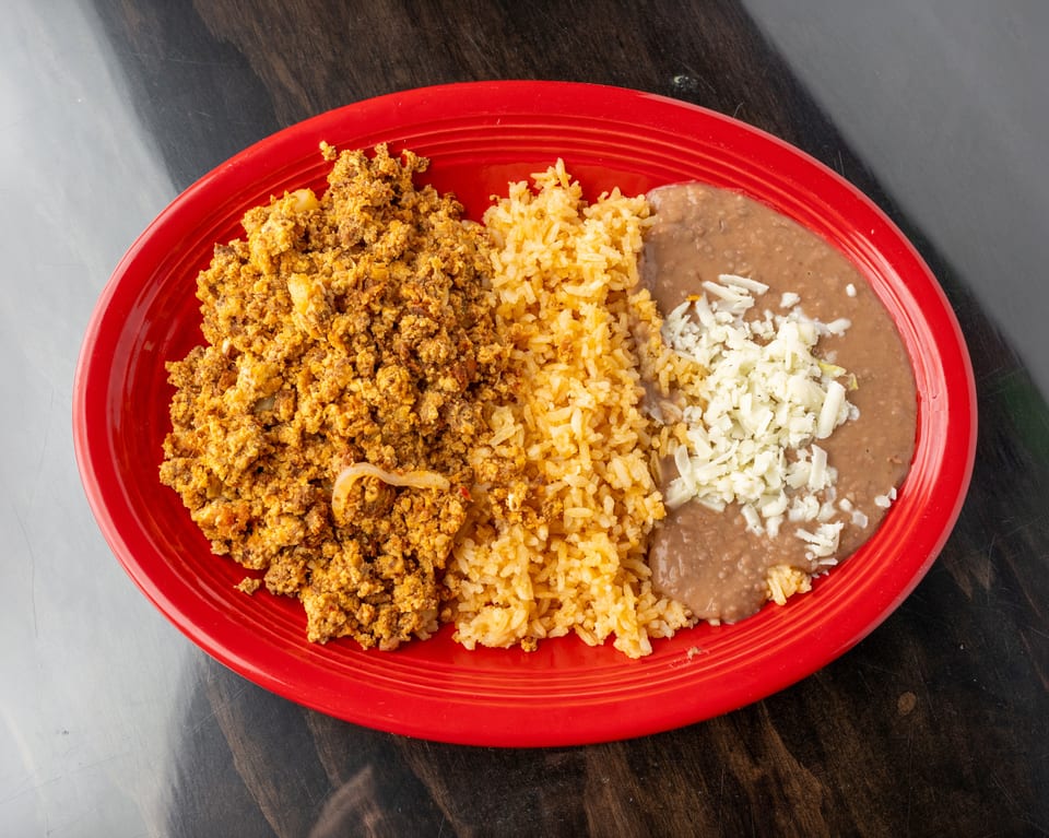 Chorizo and eggs, rice, and refried beans with shredded cheese on a red oval plate.